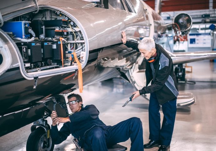 Elderly female aircraft mechanic helping her colleague who's working on the airplane's landing gear retraction system. She is passing him a tool.